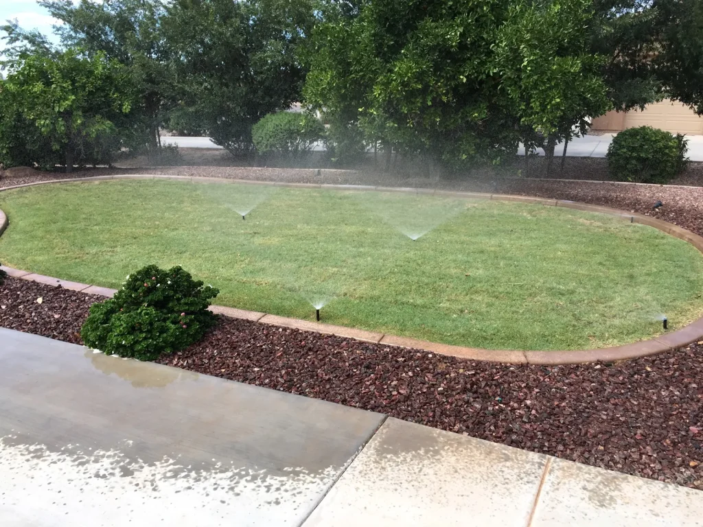 Sprinklers watering an oval grass lawn edged with red decorative rock
